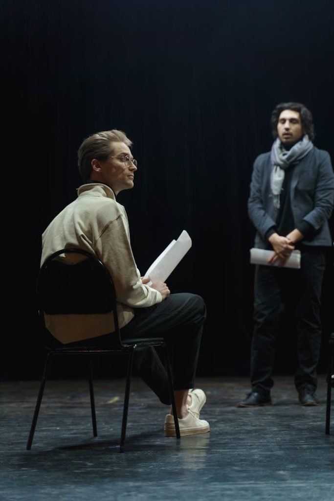 Two actors engage in a rehearsal, one seated and the other standing, holding scripts on a theater stage.